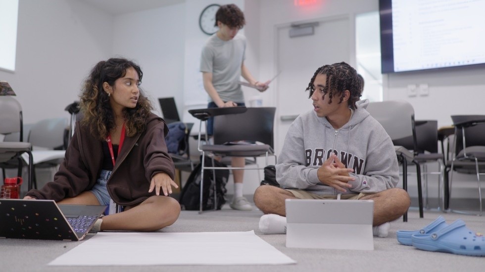 Students sitting on the classroom floor discussing their work with laptops