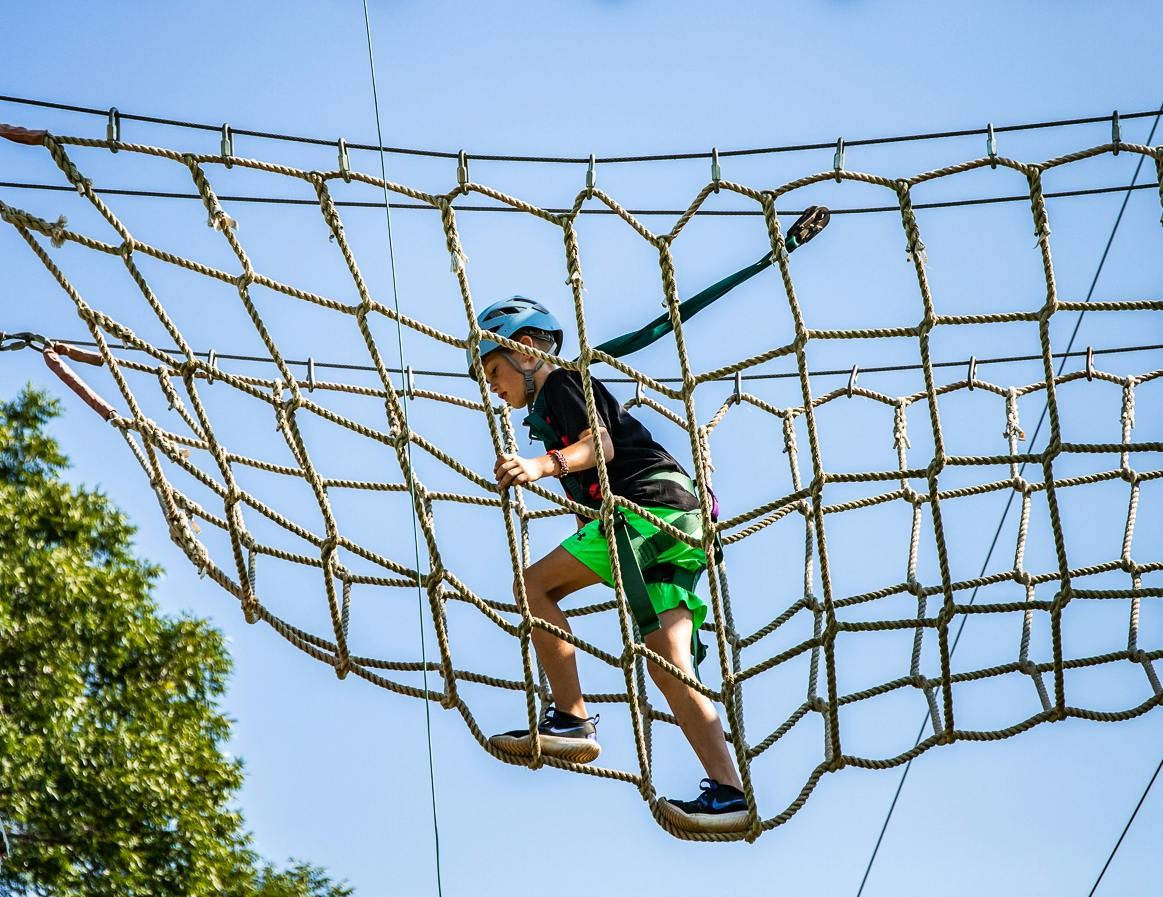A helmeted camper crosses an elevated rope-net bridge