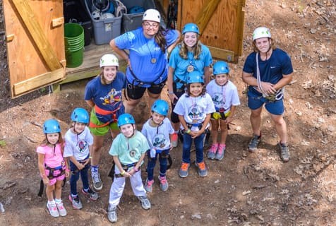 Helmeted campers and staff in harnesses gather beside a ropes-course structure