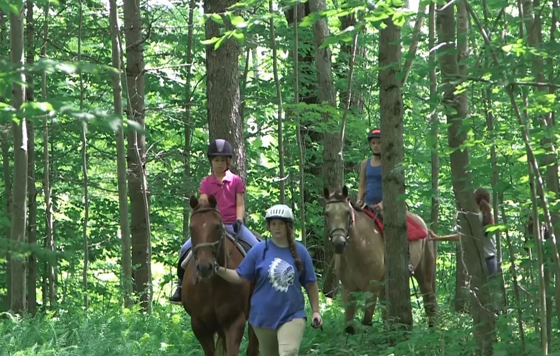 A camper riding a horse with a staff member in the woods