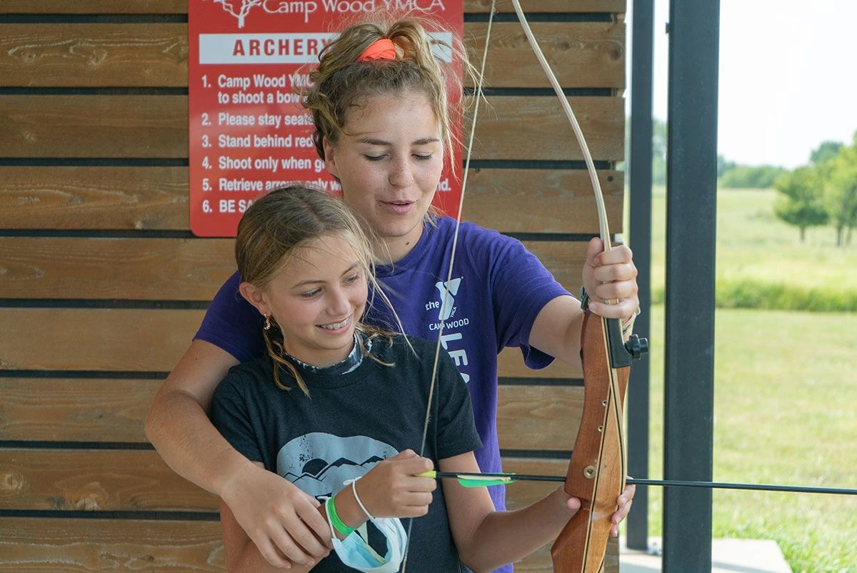 A counselor helps a camper hold a bow at the archery range