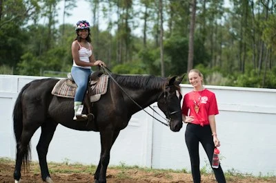 A helmeted camper sits on a horse beside a staff member