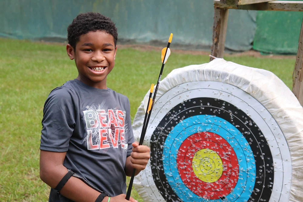 A camper stands with archery equipment beside a target